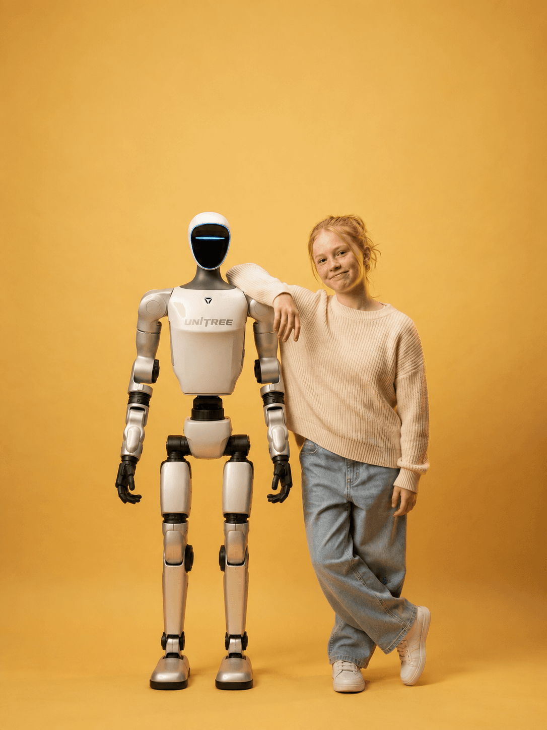 Unitree G1 humanoid standing beside a person against a warm amber backdrop